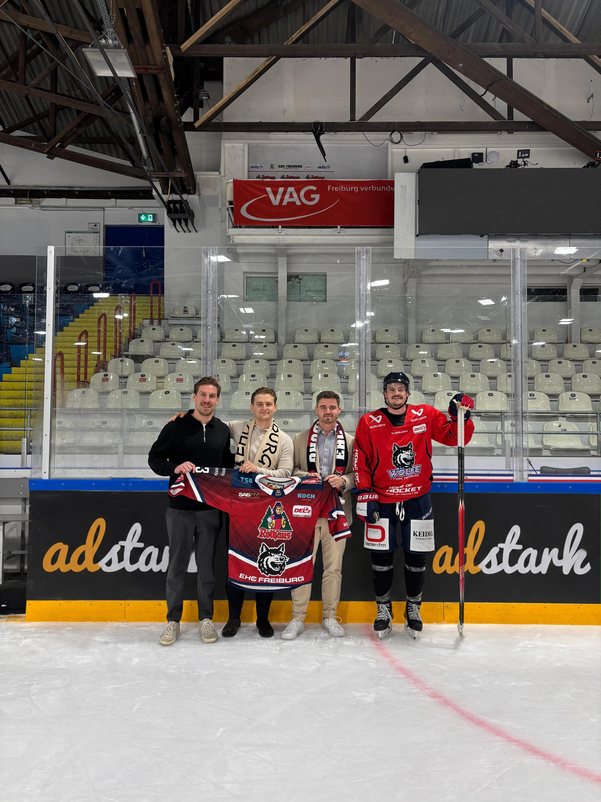 Das Adstark®-Team mit Trikot und Schal des EHC Freiburg und einem Spieler auf dem Eis, dahinter die Bande mit dem Adstark®-Logo